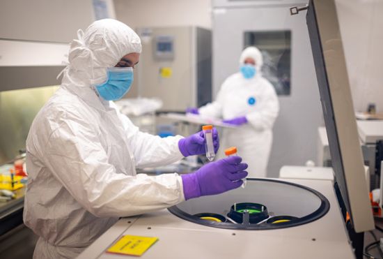 Person wearing full surgical dressing holding two vile tubes with person in background wearing surgical dressing